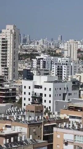 The video displays a panoramic daytime view of a dense urban environment, captured from an elevated perspective, likely a high-rise building or rooftop. The camera slowly pans from right to left across the cityscape.

In the foreground and midground, numerous multi-story residential and commercial buildings are visible, predominantly in shades of white, beige, and light grey. Many rooftops are equipped with an abundance of solar water heaters and air conditioning units. Trees and other vegetation are interspersed among the buildings.

In the distant background, a prominent modern city skyline stretches across the horizon, featuring numerous tall skyscrapers and several visible construction cranes, indicating ongoing urban development. The architectural style and the specific configuration of the distant high-rises strongly resemble the skyline of Tel Aviv, Israel.

The weather is clear and sunny, with a bright blue sky. There are no people visible in the video, nor is there any discernible text.