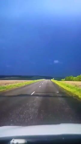 The video shows a POV of a vehicle driving on an asphalt road. The road has white dashed lines indicating lanes. The landscape on either side of the road consists of green fields and trees. The sky is a dark, stormy blue. The camera appears to be mounted on the dashboard of the vehicle. There are no people visible in the video.