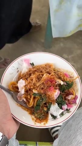 A close-up overhead shot shows a person holding a white bowl of food. The bowl contains noodles with a reddish-brown sauce, topped with shredded meat, green vegetables, and pink and white crackers. A fork is visible in the left side of the bowl, partially submerged in the food. The background is blurred, but appears to be an outdoor setting with people and a table covered with a white cloth. The lighting is natural and appears to be daytime.