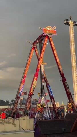 A towering amusement park ride with large red triangular supports and gold trim is the main focus of the video. The ride is illuminated with flashing pink and orange lights. At the top of the ride, a sign displays the word "DACHA" in gold lettering against a red background. In the background, a taller, thinner structure also lit with lights rises into the sky, and other carnival attractions with lights and signage are visible. The sky is overcast and dark gray, suggesting potential rain or storm clouds. People are present at the base of the ride, but their faces are not clearly visible. The ride appears to be in motion, with parts of it swinging or rotating. The overall scene depicts a bustling amusement park atmosphere.