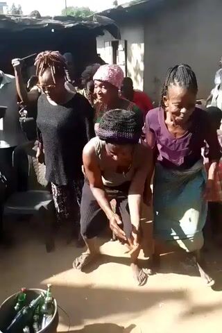 In a sunny, outdoor setting, a group of adults and children are gathered together. The adults are moving around, with some appearing to dance or participate in a celebration. The people are dressed in casual clothing, with some individuals wearing traditional African attire. A bucket filled with glass bottles sits on the ground, and some people are holding red cardboard boxes. The scene suggests a social gathering or event taking place.
