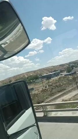 A video taken from inside a vehicle shows a landscape view from a moving vehicle. The vehicle appears to be a truck, as evidenced by the side mirror and part of the cab visible in the lower left portion of the frame. Outside the vehicle, the scene depicts a semi-arid environment with a few buildings and some debris scattered around. There are power lines and a railway track visible in the midground. The sky is blue with scattered white clouds. The terrain appears somewhat hilly and dry, with sparse vegetation. The overall impression is of a rural or industrial area.