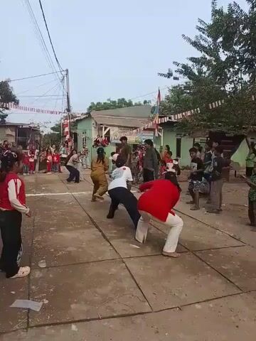 This is a video of a tug-of-war game in what appears to be a village or community setting. Several people are participating in the game, pulling on a rope. One team is wearing red and white, while the other team is wearing yellow and white. Spectators are gathered on the sides, watching the game. The background shows houses and trees, with Indonesian flags and decorations strung up, suggesting a celebration or event. The ground is paved with square tiles. The overall atmosphere seems lively and festive.