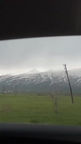 The video shows a view from inside a moving vehicle. The camera is pointed towards the outside, capturing a landscape scene.  The foreground features a green field interspersed with power lines and wooden poles.  In the middle ground, there are several leafless trees.  The background reveals a mountain range partially covered in snow under an overcast sky. The vehicle appears to be traveling on a road parallel to the field. The framing of the video suggests it's taken from the passenger side window of the car. The overall impression is one of a rural setting on a cloudy day.