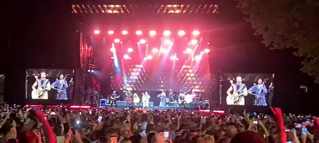 This is a wide shot of a concert crowd at night. The stage is illuminated with bright pink and white lights, and there are large screens on either side displaying live footage of the performers. A band is playing on stage, with several musicians visible, including guitarists, a drummer, and vocalists. The crowd is densely packed, with many people holding up their phones to record the performance. Some audience members are wearing red accessories. The overall atmosphere appears to be energetic and celebratory.