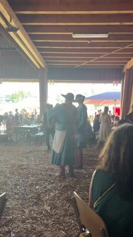 The video shows an indoor space with a wooden roof structure and corrugated metal roofing. The floor is covered in wood chips. In the foreground, chairs are visible. In the background, a gathering of people is taking place. Several individuals are dressed in what appear to be period costumes, with one person in a teal tunic and apron, and another in a blue tunic and hat. Others are in more modern attire. Some people are standing and interacting, while others are seated at tables. The lighting suggests it is daytime, with sunlight entering the space from open sides. There are also some decorations or banners hanging from the ceiling. The overall atmosphere appears to be that of a historical reenactment or a themed event.