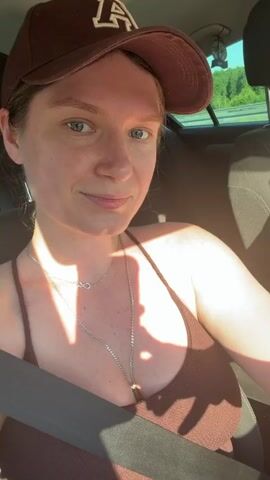 A woman wearing a brown baseball cap and a brown tank top is sitting in the passenger seat of a car. She is smiling and looking at the camera. Sunlight is casting shadows on her chest. A man wearing a green t-shirt and a black baseball cap is driving the car. He is looking out the window. The car appears to be on a highway, with trees and sky visible in the background.