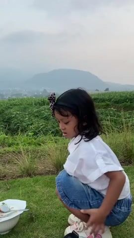 A young girl with a black bow in her hair is squatting down on the grass in front of a plate of food. She is wearing a white shirt and denim shorts. The background is a green landscape with mountains in the distance. She then stands up and walks away from the camera.
