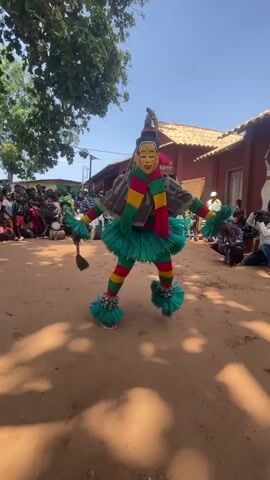 A person in a colorful costume with a mask dances in the center of a dirt area. The costume is primarily green, red, and yellow, with fluffy pom-poms on the feet and wrists. The mask has a yellow face with red lips and green eyes. The dancer has their arms outstretched. In the background, a crowd of people are gathered, some seated and some standing. There are buildings with red walls and tile roofs behind the crowd. Trees are visible on the left side of the frame. The sky is clear and blue. The ground is a reddish-brown dirt. There are shadows on the ground, suggesting it is daytime.