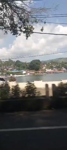 The video shows a view from a moving vehicle looking out at a coastal town. The scene is dominated by a body of water, likely a bay or sea, with buildings and greenery on the hills in the background. In the foreground, there are some parked vehicles, including white vans and a black van, on a paved area. There are also some trees and foliage. The sky is partly cloudy. The video appears to be shot during the daytime.