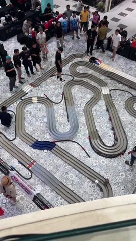 A high-angle shot shows a Mini 4WD track laid out on a patterned floor in a public space, likely a mall. Several people stand around the track, watching the cars race. The track is made of grey plastic segments forming loops, curves, and straightaways, with some sections elevated. Two toy race cars, one red and one blue, are actively racing around the track at high speed. Spectators stand around the track, some looking down at the cars, others conversing. A few individuals are seated or crouching near the track, possibly making adjustments. Display banners for "Tamiya Mini 4WD" are visible near the front of the frame. The environment suggests an organized event or competition taking place in a shopping mall or similar commercial space.