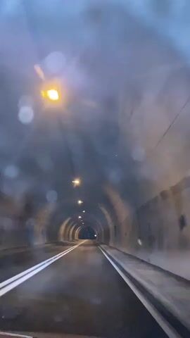This is a first-person view video of a car driving through a tunnel. The tunnel is made of concrete and has lights installed along the ceiling. The road surface is wet, and there are white lines marking the lanes. The tunnel appears to be somewhat foggy or misty, with light scattering from the lamps. The camera is mounted inside the car, showing the dashboard and the front windshield. The video captures the car moving forward into the distance, towards the brighter opening of the tunnel.