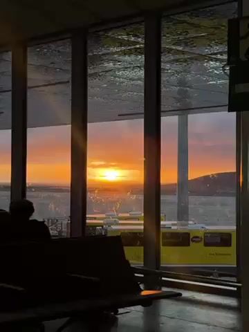 The video shows a sunset as seen through a window in an airport or similar waiting area. The sun is a bright orange, low on the horizon, and the sky is filled with shades of orange, pink, and purple. The window is composed of multiple panes, with condensation on the glass. In the foreground, there are rows of empty seats, silhouetted against the light. To the right, a yellow bus is visible outside. A person is seated on one of the seats, also in silhouette.