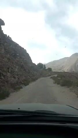 The video shows a view from inside a vehicle, likely a car, as it travels on a dirt road through a mountainous, arid landscape. The road is unpaved and appears to be winding uphill. On the left side of the road, there is a steep, rocky mountainside. Sparse vegetation, including some shrubs and a few trees, is visible on the sides of the road and in the distance. The sky is overcast with clouds. The interior of the vehicle is visible in the foreground, including the dashboard and windshield. The video is shot from a fixed perspective within the vehicle, capturing the motion of the car as it navigates the terrain.