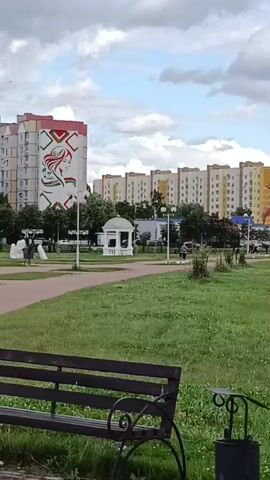 The video shows a park with a walking path and benches. In the background, there are apartment buildings and a gazebo. There are a few people walking in the park and some sculptures. The sky is cloudy.