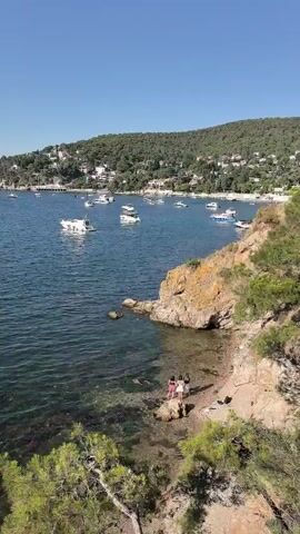 The video shows a scenic view of a bay with boats and a coastline. In the foreground, there is a rocky area with some greenery. Two people are visible on the rocks near the water. The bay is filled with several boats of various sizes, including yachts and smaller vessels. In the background, a green, tree-covered hill slopes down to the water, with buildings scattered along the hillside. The sky is clear and blue, indicating a sunny day. The water appears clear and blue-green, with some aquatic plants visible near the shore. The overall scene depicts a peaceful coastal environment with recreational boating activity.