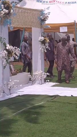 The video depicts a funeral service held outdoors on a grassy area. A white coffin, adorned with gold-colored hardware, is positioned on a raised platform decorated with white fabrics and artificial flowers. The platform is framed by white pillars. People are walking around, some dressed in traditional attire. A photographer is taking pictures of the scene. A white tent is visible in the background.