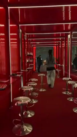 A person with long dark hair walks down a long red corridor. The corridor is lined with small silver swivel stools and red counters. The walls, ceiling, and floor are red. There are bright white lights on the ceiling along the edges of the space. There are two other people at the far end of the corridor. One is wearing a dark coat and the other is wearing a white coat.
