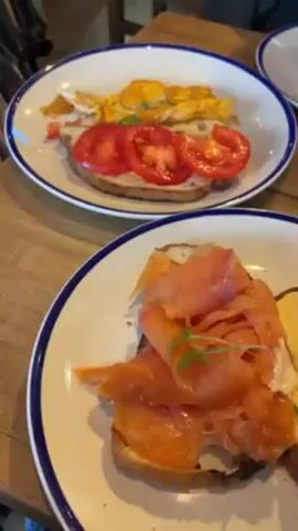 The video shows three plates of food on a wooden table, likely in a restaurant. The plates are white with a blue rim. In the foreground is a plate with two halves of toast, one topped with salmon and the other with two poached eggs covered in hollandaise sauce. Microgreens garnish both.  The middle plate has toast topped with sliced tomatoes and some scrambled eggs.  The third plate has avocado toast topped with two fried eggs and bacon. A spoon and bottle of red liquid are also visible on the table. Someone's hand, holding a knife, moves in the background.  The scene focuses on the food, showcasing a selection of breakfast or brunch dishes.