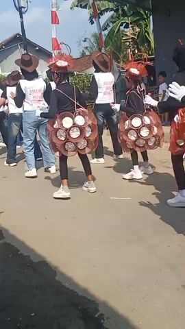 The video shows a group of people walking in a parade on a paved road. Several people in the foreground are wearing costumes. Two individuals in the middle ground have circular, cage-like skirts made of brown material and white cylindrical objects. They are wearing black tops and hats. To the right, another person is wearing a red and white polka-dotted skirt and a black top, with a black hijab. Several other individuals in the background are also walking in the parade, some wearing white vests with text on them and cowboy hats. A red and white flag is visible in the background, along with houses and trees. The weather appears to be sunny.
