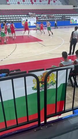 The video captures a junior basketball game in an indoor arena. The court is made of polished wood with distinct red and white markings. Spectators are seated in tiered red and white bleachers, some visible in the foreground and background. A Bulgarian flag is prominently displayed in the foreground, draped over a railing.

On the court, young athletes in red and green uniforms are engaged in a basketball game. Several players are actively participating in the play, including dribbling, passing, and shooting. One player in a green jersey with the number 13 is in focus in the foreground. The scoreboard in the background shows the time as 5:11.

The setting appears to be a modern sports facility, likely in Çanakkale Merkez, Turkey, as indicated by the flags and event banners. The overall atmosphere seems to be one of a competitive sporting event, with young athletes focused on the game. The lighting suggests it is daytime, as the arena is well-lit. The visible text includes "FIBA YOUTH DEVELOPMENT" on a banner, and potentially national flags of participating countries hanging from the ceiling. The emotion conveyed is one of focused athletic performance.