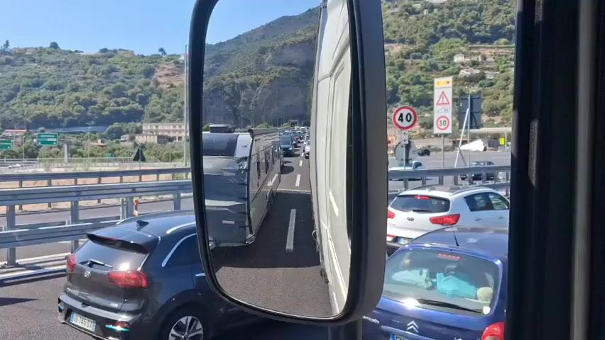 The video shows a view from a vehicle, likely a truck, stuck in a traffic jam. The camera is positioned to capture the side mirror and the road ahead. The side mirror reflects a grey caravan being towed, with other vehicles visible behind it. The main view shows a long line of cars and trucks at a toll plaza on a sunny day. Green, hilly terrain with some buildings is visible in the background. Traffic signs indicating a speed limit of 40 are also visible. The traffic appears to be stopped or moving very slowly.