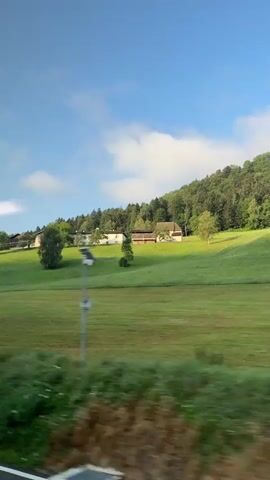 The video shows a scenic view of a rural landscape with rolling green hills. There are several houses and farm buildings nestled on the hillsides, surrounded by trees. The sky is a clear blue with a few scattered white clouds. The video appears to be taken from a moving vehicle, as evidenced by the slight blur of the foreground and the continuous movement of the landscape. The overall impression is one of tranquility and natural beauty.