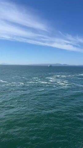 A wide shot of the ocean on a clear day with a bright blue sky and scattered white clouds. A distant white boat is visible on the horizon. The water is choppy with whitecaps. The perspective is from a moving vessel, as evidenced by the wake in the foreground. In the latter part of the video, a ferry terminal comes into view on the right side of the screen, with structures and boats docked. The sun is shining brightly, reflecting off the water.
