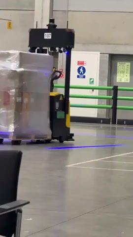 A black and yellow autonomous mobile robot, designated by the number 107 on its chassis, is transporting a pallet wrapped in clear plastic shrink wrap. The robot has blue lights projecting onto the floor around its base, indicating its operational status. In the background, another autonomous mobile robot, also black and yellow, is visible to the left of the primary robot. The robots are operating within a large, industrial warehouse environment. The floor is concrete, marked with white lines that appear to designate pathways or operational zones. A green barrier and yellow safety cones are also present in the background, along with a large roll-up door. The lighting is typical of an indoor industrial setting.