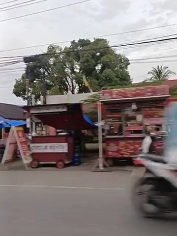The video shows a street scene with several food stalls and vehicles. On the left, there is a blue tent with a person standing behind a counter. Next to it is another blue tent with a food cart. In the middle of the street, a white car drives by. Further down the street, there are more food stalls, some with people around them. A motorcycle is seen on the left side of the screen. There are power lines overhead and trees in the background. The scene appears to be in a relatively urban or suburban area, possibly in Southeast Asia, given the architecture and the presence of food carts. The overall atmosphere is bustling with activity.