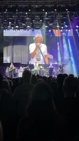 The video shows a live concert performance from the perspective of the audience. A large screen behind the performers displays a close-up of a man singing into a microphone, wearing a white t-shirt. The man on the screen appears to be an older gentleman with graying hair. On the stage below the screen, a band is performing, with multiple musicians visible playing instruments such as guitars and drums. The stage is lit with bright spotlights, primarily in shades of blue and purple, creating a dramatic effect. In the foreground, the silhouettes of audience members are visible, their backs to the camera, facing the stage. The overall atmosphere suggests an energetic and engaging musical event.