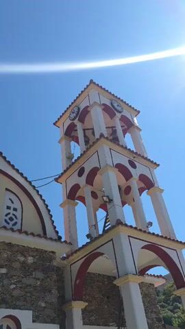 A low-angle shot shows the side of a church with a bell tower against a bright blue sky. The church has a stone facade and a curved roof tiled in red. Above the stone, the building is painted yellow with white arched trim and ornate windows. The bell tower is white with red arches and trim. It has clocks on two sides and bells hanging within. The sun is shining brightly, creating lens flare in the upper left corner.