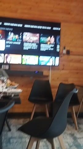 The video shows a room with wooden walls and ceiling. A large television is mounted on the left wall, displaying a menu interface. Below the television, there is a soundbar and a shelf. Two black chairs with wooden legs are placed in front of the television. To the right of the chairs is a small round table with a black top and wooden legs. On the table, there are a bottle of water, a vase with flowers, a candle, and another bottle. A large window with sheer curtains is visible on the right side of the room, with greenery and trees seen outside. The floor is covered with a patterned rug.