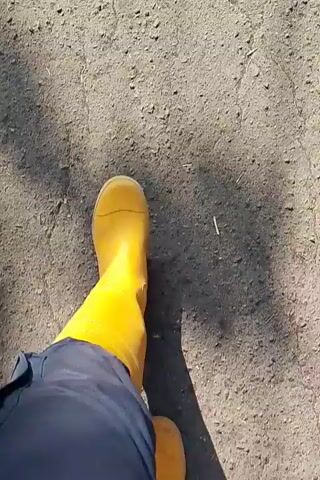 The video shows a first-person perspective of someone walking on a gravel path. The person is wearing yellow boots and dark pants. The path is rough and uneven, with some cracks visible. Sunlight casts shadows on the path.