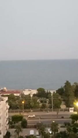 The video pans across a coastal town. In the foreground, a white apartment building with balconies is visible on the left. Beyond that, a street with traffic is visible. Cars and lights are present on the street. Trees and other buildings are scattered throughout the town. In the background, the calm sea stretches to the horizon under a pale sky.