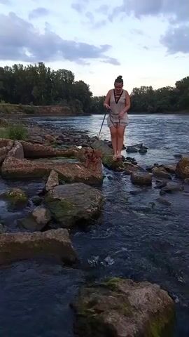 A woman with dark hair in a bun, wearing a light-colored halter top and shorts, stands on a rocky embankment next to a river. She holds a leash attached to a small brown dog that is standing on a rock to her left. The woman appears to be talking on her phone and looking down. The river is wide with a moderate current, and trees line the opposite bank. There are also some broken concrete structures in the water near the woman. The sky is overcast.