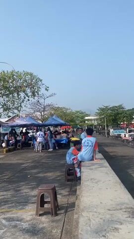 A vertical video shows an outdoor scene with a bright, clear blue sky. In the foreground, a concrete walkway slopes down slightly from a raised concrete barrier. Two plastic stools are visible on the walkway, one closer to the camera. Two young males, wearing blue t-shirts with red trim and lettering on the back, are sitting on the raised barrier. The one on the left is sitting on a plastic stool placed on the barrier, and the one on the right is sitting directly on the barrier. They are facing away from the camera, looking towards the right side of the frame. Their t-shirts have text that appears to read "SMA NEGERI 6 MANADO".

In the middle ground, a group of people are gathered under several large, colorful tents. These appear to be market stalls. There are tables set up, and people are standing and sitting around them. Some individuals are wearing white t-shirts with numbers, such as "22". Cars and possibly other vehicles are parked behind the stalls on the left side of the frame. Trees with green foliage are interspersed throughout this area.

The background shows more trees and buildings. The overall impression is a lively outdoor gathering, possibly a market or event, taking place on a sunny day near a body of water, suggested by the boats visible on the far right of the frame. The lighting indicates it is daytime, likely morning or afternoon.