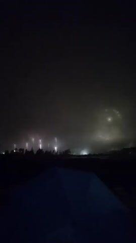 This video captures a nighttime fireworks display. The sky is illuminated with bright pink and red lights, indicative of the explosions. In the foreground, a dark, silhouetted tent is visible, suggesting the viewpoint of someone observing the event. The landscape appears to be a flat expanse, possibly a field or a body of water, with a line of trees or structures forming a dark silhouette against the illuminated sky. The fireworks are concentrated in the distance, with some appearing as bursts of light and others as glowing spheres against the smoky, colorful atmosphere. The overall impression is one of a vibrant and expansive pyrotechnic show against a dark, hazy night.