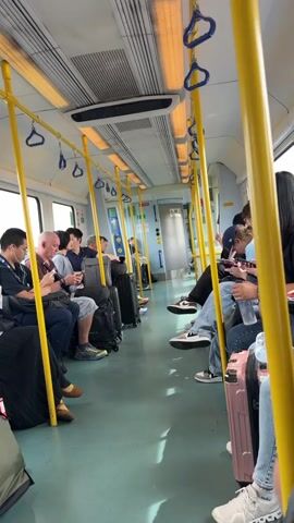 The video shows the interior of a train carriage with several passengers. Most of the passengers are seated, and many have luggage with them. The seats are arranged facing each other, with an aisle in between. Yellow poles and handrails are visible throughout the carriage. Some passengers are looking at their phones. The lighting appears to be artificial, coming from overhead fixtures. The overall impression is of a commuter or intercity train in motion.