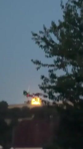 The video shows a large, glowing orb, likely the sun or moon, setting behind a distant treeline. A pole-like object with a red light at its tip is visible in front of the orb, moving upwards and to the left. The foreground shows the rooftops of houses and trees. The sky is a dusky blue, indicating either dawn or dusk.