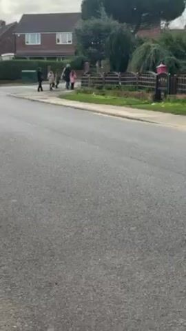 The video shows a street scene on an overcast day. A group of people, including adults and children, are walking on the opposite side of the street from the camera. They appear to be walking away from the camera.  Houses with brick facades and green hedges line the street. A green trash bin is visible near the group. The road is paved and slightly wet in appearance.