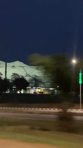 The video shows a glimpse of a large, white structure at night. The structure appears to be made of fabric stretched over a frame. Streetlights and other lights illuminate the structure, which is partially obscured by trees. There is a road in the foreground with cars passing by. The video appears to be taken from a moving vehicle. The sky is dark blue.