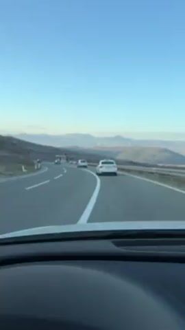 The video is shot from the perspective of the driver of a white car, looking through the windshield at a highway with light traffic. There are several cars ahead, traveling in the same direction. The road curves gently to the left. There is a green, grassy hillside to the left of the road and a guardrail to the right. Beyond the guardrail, a distant landscape of hills and mountains is visible against a clear blue sky. The video is somewhat shaky.