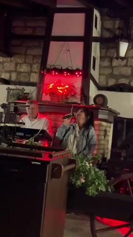 A man plays a keyboard to his left, while a woman to his right sings into a microphone. They are performing on a stage in front of a stone wall, with decorative lights in a pot of flowers above and behind them. The lights change color from green to blue to red to purple. The man is wearing a white shirt, and the woman is wearing a denim jacket over a light-colored outfit. There are speakers on the right side of the stage, and a wooden cart in the foreground.