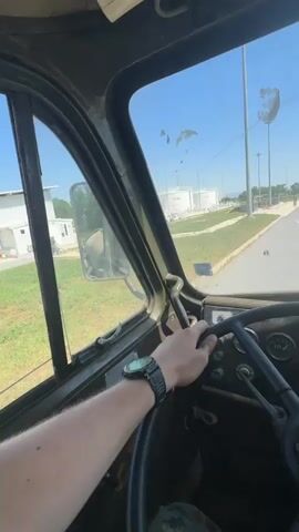 The video provides a first-person perspective from the driver's seat of a vehicle. The driver's left hand, wearing a watch, is on the steering wheel. In the background, there is a road with white lines, large white cylindrical tanks, and tall light poles under a clear blue sky. The interior of the vehicle appears to be older, with visible gauges on the dashboard. The driver is wearing camouflage pants.