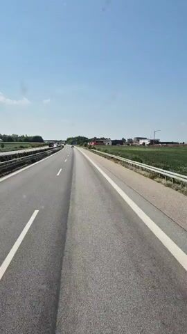 The video shows a first-person perspective of driving on a highway on a clear day. The road is paved with asphalt and has lane markings. There are guardrails along the side of the road, and on the right side, there is a field with crops. In the distance, there are some buildings and a crane. There are other vehicles on the highway, but they are far away. The sky is blue with a few white clouds.
