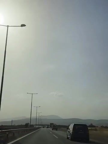 The video shows a view from a car driving on a highway. The sky is bright and hazy, suggesting a sunny but possibly polluted or dusty day. Several streetlights line the left side of the highway, their lamps illuminated. In the distance, mountains are visible. Other vehicles, including cars and trucks, are also on the highway, moving in the same direction. The perspective is from the front passenger seat, looking forward. The overall impression is one of a typical highway journey on a bright day.