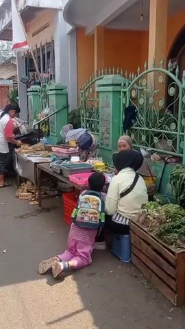 A group of people are gathered near a market stall. A child is kneeling on the ground in front of the stall, facing away from the camera. Several women are standing and interacting with the stall. The stall is covered with various goods, including food items and produce. A fence and a building are visible in the background. The scene appears to be outdoors, with sunlight casting shadows on the ground.