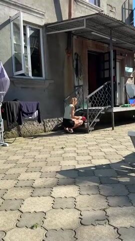 In Zugdidi, Georgia, a young woman with blonde hair tied in a ponytail is seen from the side, wearing a light green sleeveless top, black shorts, and red gloves. She is kneeling on a textured stone patio, next to a metal staircase. She is actively painting the metal steps with a brush, holding a white can of paint with orange lettering that reads "PEN" and appears to have more text below it. Another green plastic bag is visible on the landing of the staircase behind her, along with an umbrella hanging on the wall of the building. The sun is casting shadows, suggesting it is daytime, likely a sunny day. The overall scene depicts a person engaged in outdoor maintenance, possibly repainting the stairs of a house. The expression on her face is not clearly discernible, but her posture suggests focus and effort. The environment appears to be residential, with a building and a patio area.
