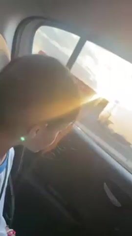 The video shows a passenger, likely a child, in the back seat of a car. The view is from behind, with the child's head partially obscuring the window. Outside the window, the sun is setting, creating a bright glow in the sky with visible clouds. The scene suggests the car is moving, likely on a highway, as indicated by the guardrail visible outside. The car interior is dark, highlighting the contrast with the bright sunset.
